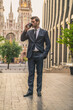 © Roman - Coffee break. Confident young man in full suit holding coffee cup and looking away while standing outdoors with cityscape in the background