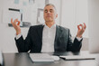 © studioprodakshn - successful gray-haired businessman wearing elegant suit sits in office holding hands in peaceful yoga position while working on business project , no stress at work concept