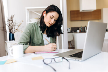 Wall Mural - Online education. A young attractive caucasian student girl writes information in her notebook during online learning at home