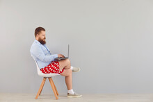 Funny Fat Man In A Blue Shirt And Red Shorts Sitting On A Chair Working Online Using A Laptop On A Gray Background.