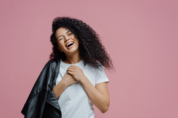 Wall Mural - Overjoyed dark skinned woman keeps hands together and laughs sincerely, closes eyes and feels very happy, shows white perfect teeth, wears t shirt and leather jacket on shoulder, isolated on pink wall