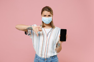 Wall Mural - Young girl in striped shirt face mask isolated on pink background. Epidemic pandemic coronavirus 2019-ncov sars covid-19 flu virus concept Hold mobile phone with blank empty screen showing thumb down.