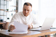 © dikushin - Business man wearing fashion casual clothing is typing on laptop computer and working with paper document at the table in modern office room on background of large window. Concept of office working.