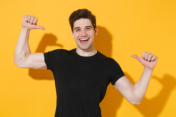 Cheerful young man guy 20s wearing casual black t-shirt posing isolated on yellow background studio portrait. People sincere emotions lifestyle concept. Mock up copy space. Pointing thumbs on himself.