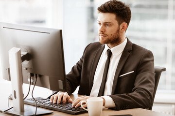 Business concept. Successful young businessman at work. Manager sitting at the office table and working on computer. Busy man in suit indoors on glass window background