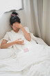 © Prathankarnpap - An ill woman holds a glass of water and consuming a pill while lying on the bed in the bedroom.