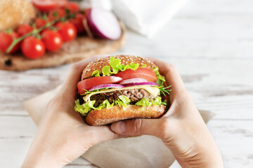 Woman hands hold beef burger with fresh salad, cheese and tomatoes over table.