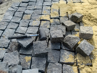 A black rubber mallet lies in the midst of natural gray granite stones and yellow sand. Cobblestone paving - tools and materials for work