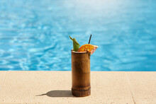 Exotic Cocktail In Wooden Glass With Lemon And Drinking Tube Standing On Poolside With Blue Pool's Water On Background, Tasty Fresh Beverage On Resort.