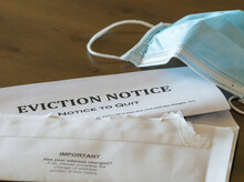 Defaulting Renter With Facemask Receives Letter Giving Notice Of Eviction From Home On Wooden Table