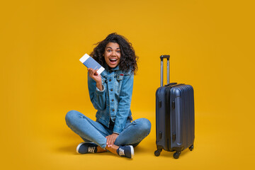 Wall Mural - Full-length portrait of smiling tourist girl sitting with her suitcase, holding passport and boarding pass and looking directly to the camera with happy face expression