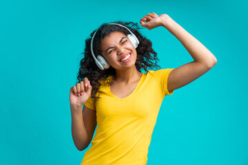 Wall Mural - Cute dark skinned girl having fun enjoying her day-off singing and dancing while listening music via wireless headset over blue background