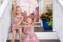Two Cute Little Girls Sitting On The Front Porch On Fourth Of July Waving Flags