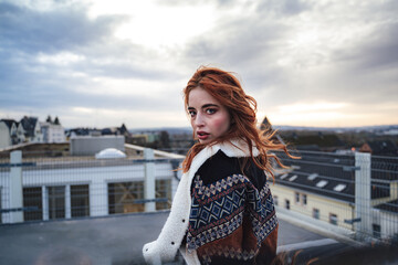 Wall Mural - Photo of beautiful girl with long red hairs wearing jacket and looking to camera. Female model standing on flat roof of building.