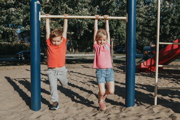 Two funny Caucasian friends hanging on pull-up bars in park on playground. Summer outdoors activity for kids. Active children boy and girl doing exercises sport. Healthy happy childhood.