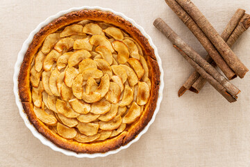 Wall Mural - Homemade baked French apple tart, an open faced apple pie, in a baking white ceramic dish aside cinnamon sticks, all on a natural linen table cloth. Flat lay, top view.