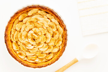 Wall Mural - Homemade baked French apple tart, an open faced apple pie, in a baking white ceramic dish aside a white cooking spoon and a white napkin on a white background. Flat lay, top view with copy space.