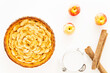 Homemade baked French apple tart, an open faced apple pie, in a baking white ceramic dish aside a white jar, Gala apples and cinnamon sticks on a white background. Flat lay, top view.