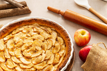 Homemade baked French apple tart, an open faced apple pie, in a baking white ceramic dish aside Gala apples in a jute bag, cinnamon sticks, cooking spoon, and rolling pin on natural linen tablecloth.