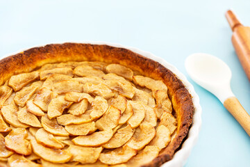 Wall Mural - Closeup on a homemade baked French apple tart, an open faced apple pie, in a baking white ceramic dish aside a cooking spoon and a rolling pin on a light blue background. Side view.