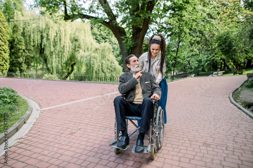 Happy young hipster modern woman with dreadlocks, walking together with her old senior grandfather, sitting in a wheelchair, outdoors in park, talking and laughing. Disabled man with his care giver