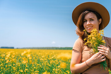 A happy beautiful young brunette woman in a hat on a blooming rapeseed field is full of joy and happiness. The concept of freedom and childhood, a lot of space for text on a yellow background