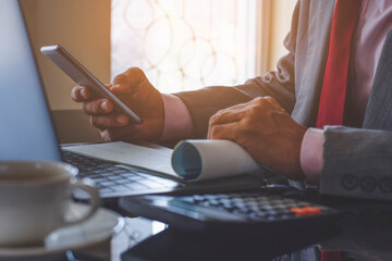 Wall Mural - Businessman in suit, hand holding and using mobile phone with checkbook, laptop computer, calculator and cup of coffee on desk at modern office. Fintech , paycheck and payroll concept .