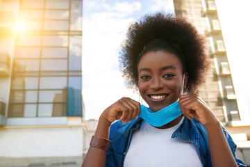 Wall Mural - Quarantine is over, coronavirus is finish. Young beautiful black african american woman with curly black hair taking her medical protective mask off. City background.