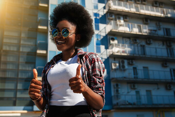 Image of a happy optimistic emotional young american african woman posing over city background dressed casual and wearing sunglasses, showing thumbs up gesture.