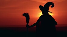 Woman In Halloween Costume Sorceress Holding Besom Waiting Posing On The Hill Sky With Yellow Sun