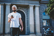 © BullRun - Handsome hispanic model dressed in casual white t-shirt with copy space for brand name or label standing on promotional background outdoors, bearded guy strolling with smartphone texting messages