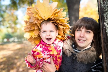 Charming young mother holds in her arms a pretty little girl with a wreath of yellow maple autumn leaves on her head while walking in the park. Autumn concept