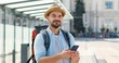 Caucasian young handsome man in hat with backpack tapping and scrolling on smartphone and smiling joyfully at bus station. Cheerful male traveller chating and texting message on mobile phone outdoor.