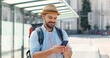 Young handsome Caucasian man in hat with backpack texting message on smartphone and smiling cheerfully at train station. Attractive male traveller tapping and scrolling on mobile phone at bus stop.