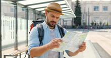 Young Handsome Caucasian Man Tourist In Hat With Backpack Holding City Map And Planning Route. Looking For Way In Downtown Train Station. Attractive Male Traveller Holding Plan Of Town At Bus Stop.
