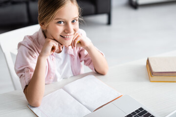 Wall Mural - High angle view of cute child smiling at camera near laptop and copy book on table