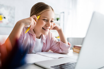 Wall Mural - Selective focus of positive kid holding pen near notebook and looking at laptop on table