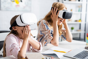 Wall Mural - Selective focus of excited kid covering mouth while using vr headsets with mother near laptop and stationery