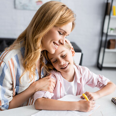 Wall Mural - Smiling mother embracing kid writing on notebook at table