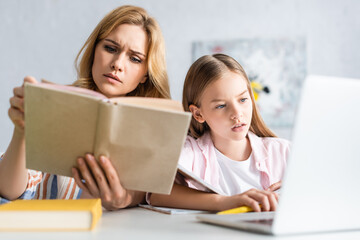 Wall Mural - Selective focus of concentrated woman reading book near daughter using laptop at table