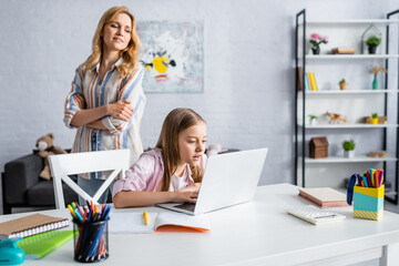 Wall Mural - Selective focus of woman looking at daughter using laptop near stationery on table