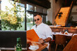 © Dragica - A middle-aged caucasian  businessman with glasses in a white shirt is sitting in a cafe, working on a laptop and holding a business folder in his hand