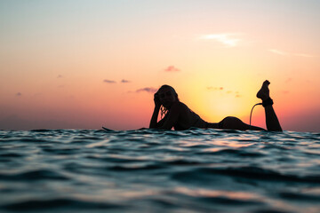 Portrait from the water of surfer girl with beautiful body on surfboard in the ocean at colourful sunset time