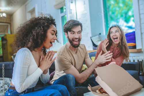 Cuadros en Lienzo Cheerful guy opening box with pizza and two enthusiastic girls