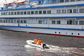 Poster - Little PVC inflatable boat with small outboard motor with two people in orange life vest overtakes a large white ferry passenger ship on a summer Sunny day, safety of boats drive on water