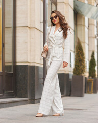 Young beautiful elegant woman in white casual costume walking at street on a summer day. Pretty lady wearing sunglasses and holding handbag. Businesswoman standing and posing in the city