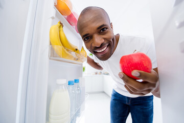 Close-up portrait of his he nice attractive cheerful cheery guy looking in fridge taking red ripe apple snack health care lifestyle regime in light white interior house kitchen indoors