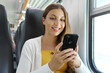 © zigres - Smiling Brazilian businesswoman using smartphone social media app while commuting to work in train. Woman sitting in transport enjoying travel.