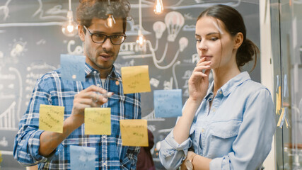Poster - Male and Female Creative Designers Create Outline of the Work Plan with Sticky Papers on Glass Wall and Draw on it with Markers. Stylish Young People in Trendy Environment.