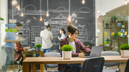 Poster - Talented Asian Developer Sits at the Table Works on a Laptop. In the Background Busy Office with Creative People Working on Personal Computersю